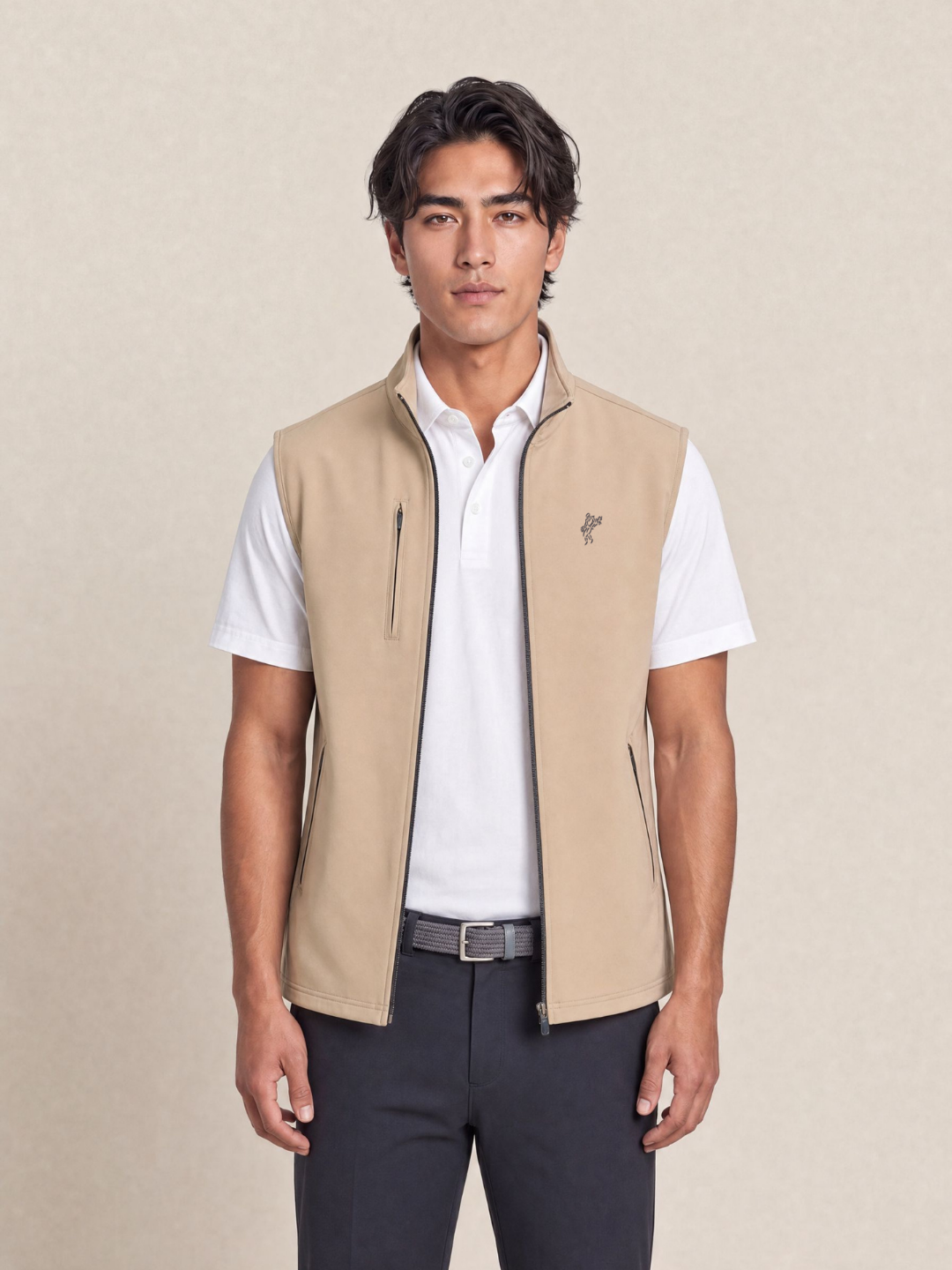 A man wearing a beige zip-front vest over a white short-sleeve polo shirt and dark trousers, standing against a plain beige studio background.