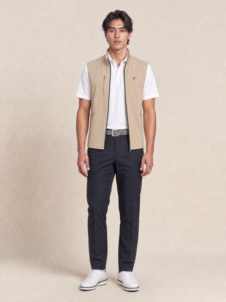 A man modeling a beige zip-front vest over a white short-sleeve polo shirt, paired with dark navy trousers and white sneakers, against a warm beige studio backdrop.